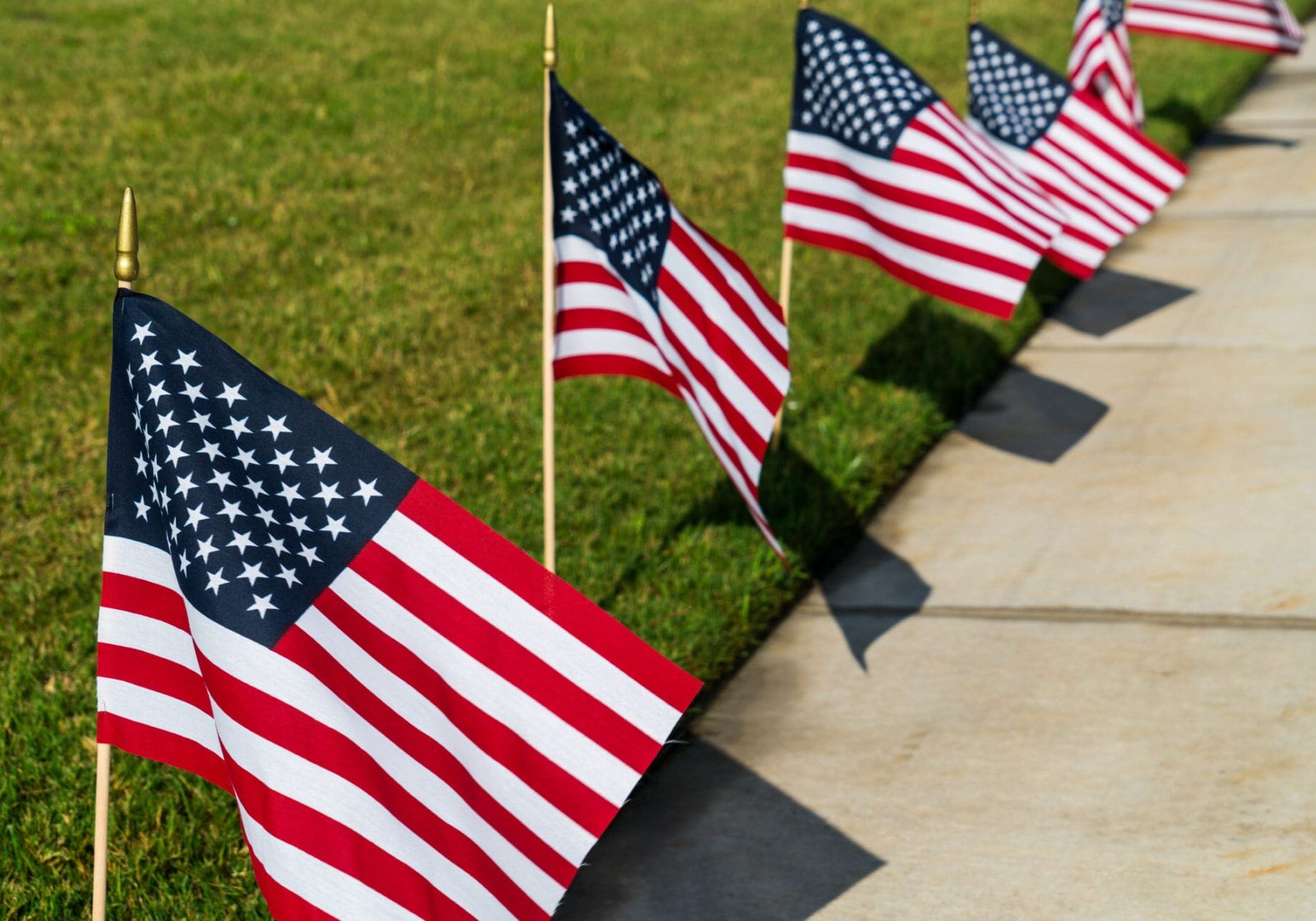 shadows of American Flags all in a Row along the sidewalk as a green grass lawn decorates for the 4th of July 2018